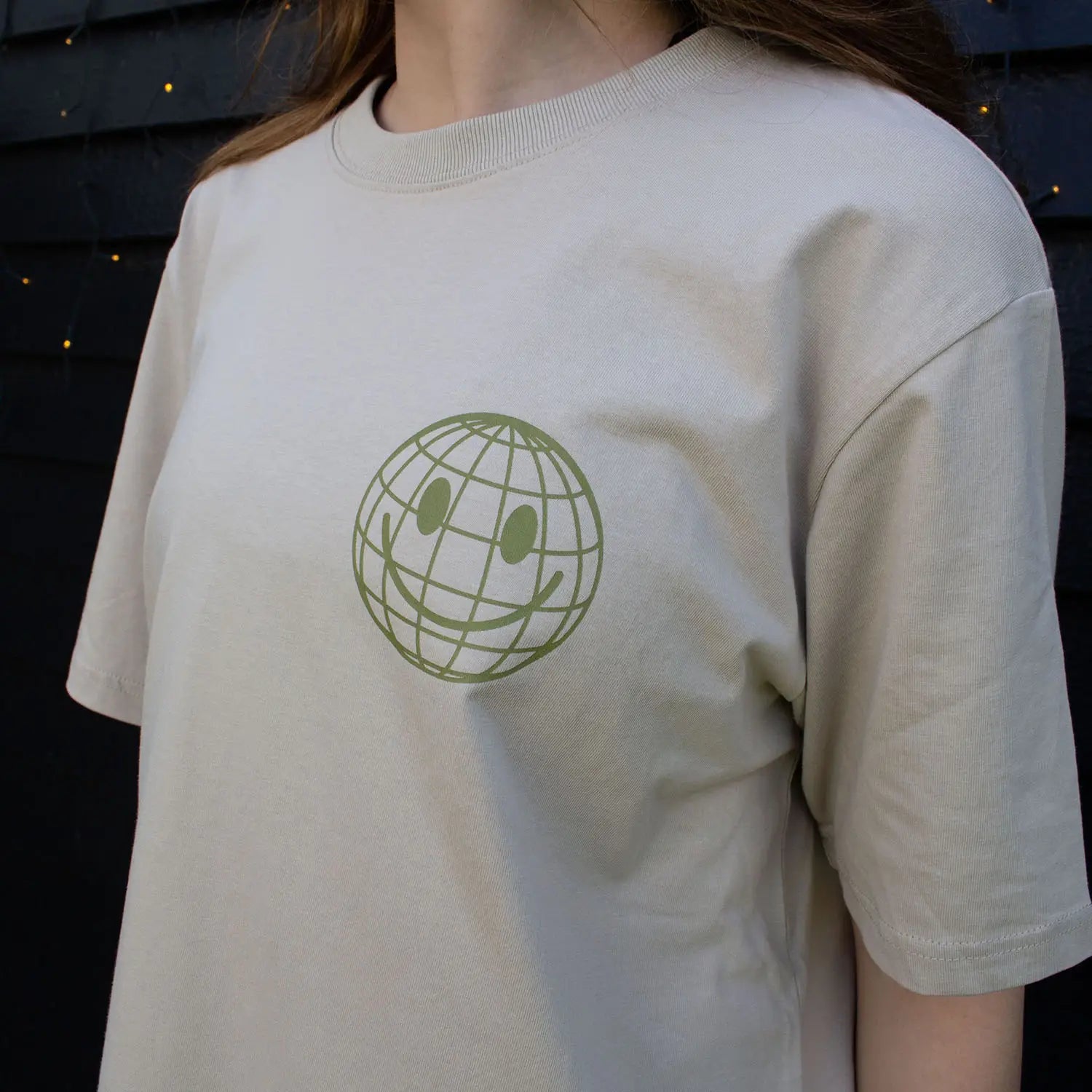 Woman wearing beige That's Sooo You t-shirt with ollive printed, smiley face design, it says Being Weird Feels Right, she is standing in front of a black wooden background with fairy lights, facing the camera.