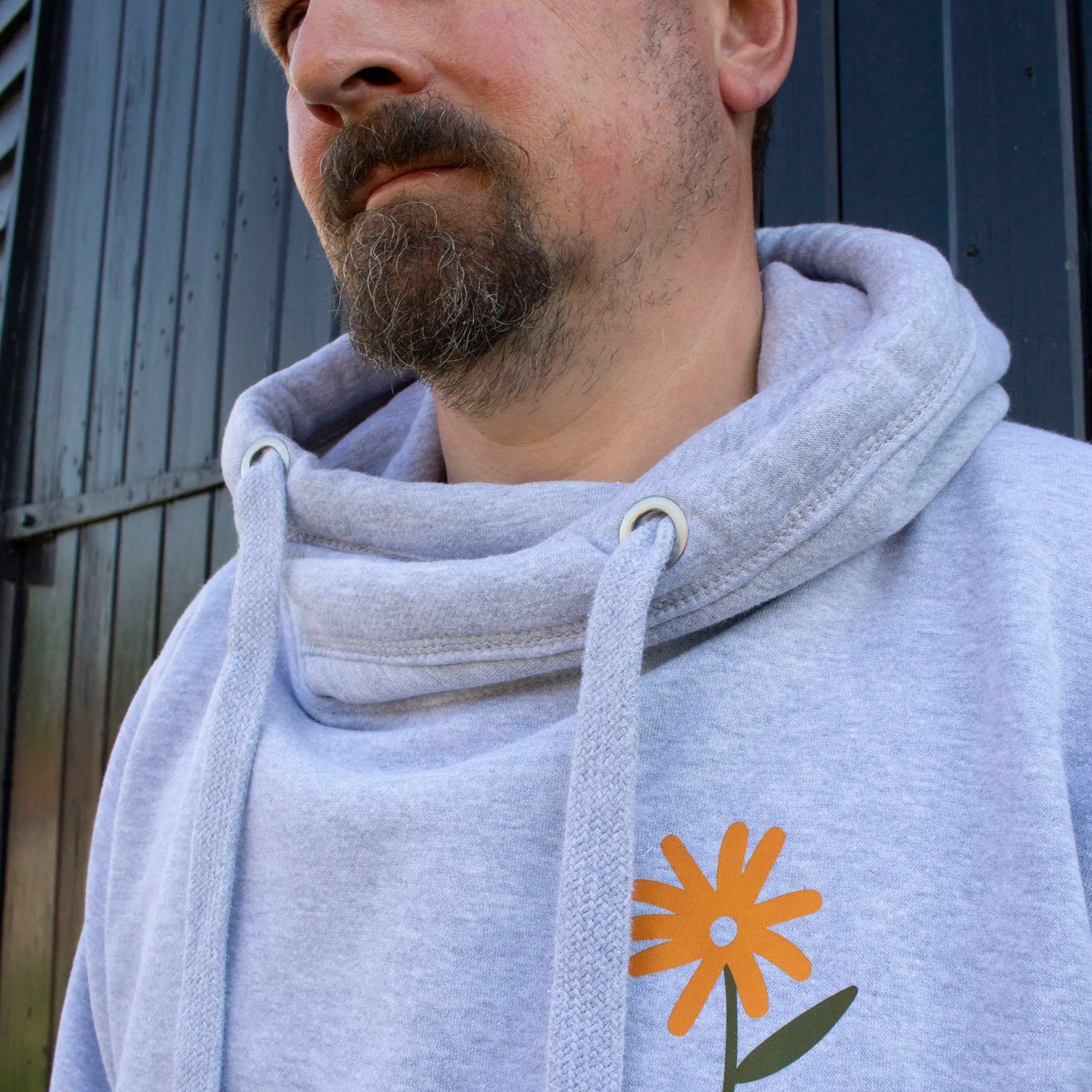 Man wearing mid-grey crossneck hoodie with chunky drawcords, That's Sooo You with orange, olive green and lilac printed, Don't Ever Stop Growing, with flowers and checkerboard design, he is standing in front of a brick background, with a TSY sign, facing the camera.