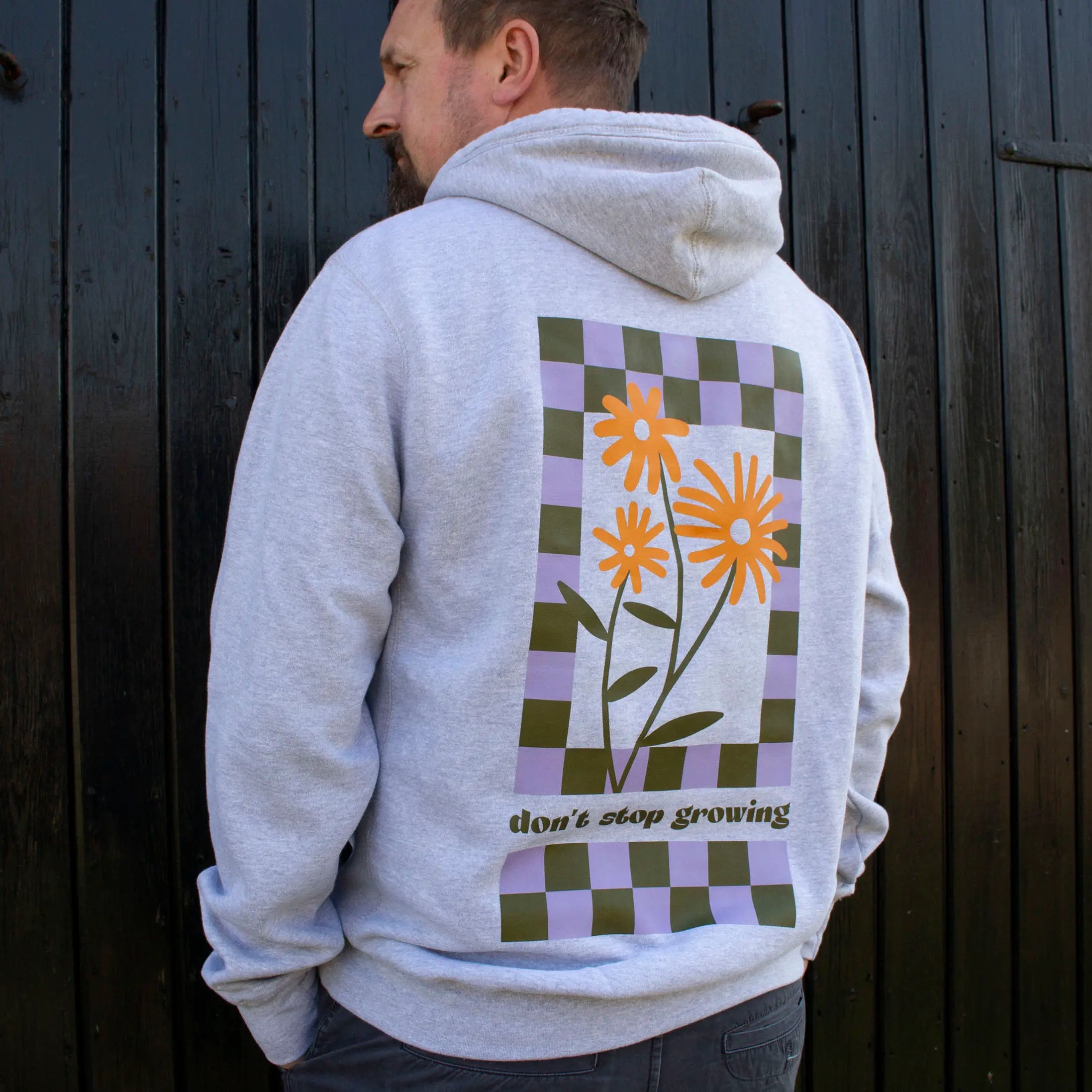 Man wearing mid-grey crossneck hoodie with chunky drawcords, That's Sooo You with orange, olive green and lilac printed, Don't Ever Stop Growing, with flowers and checkerboard design, he is standing in front of a black wooden background, facing away from the camera.
