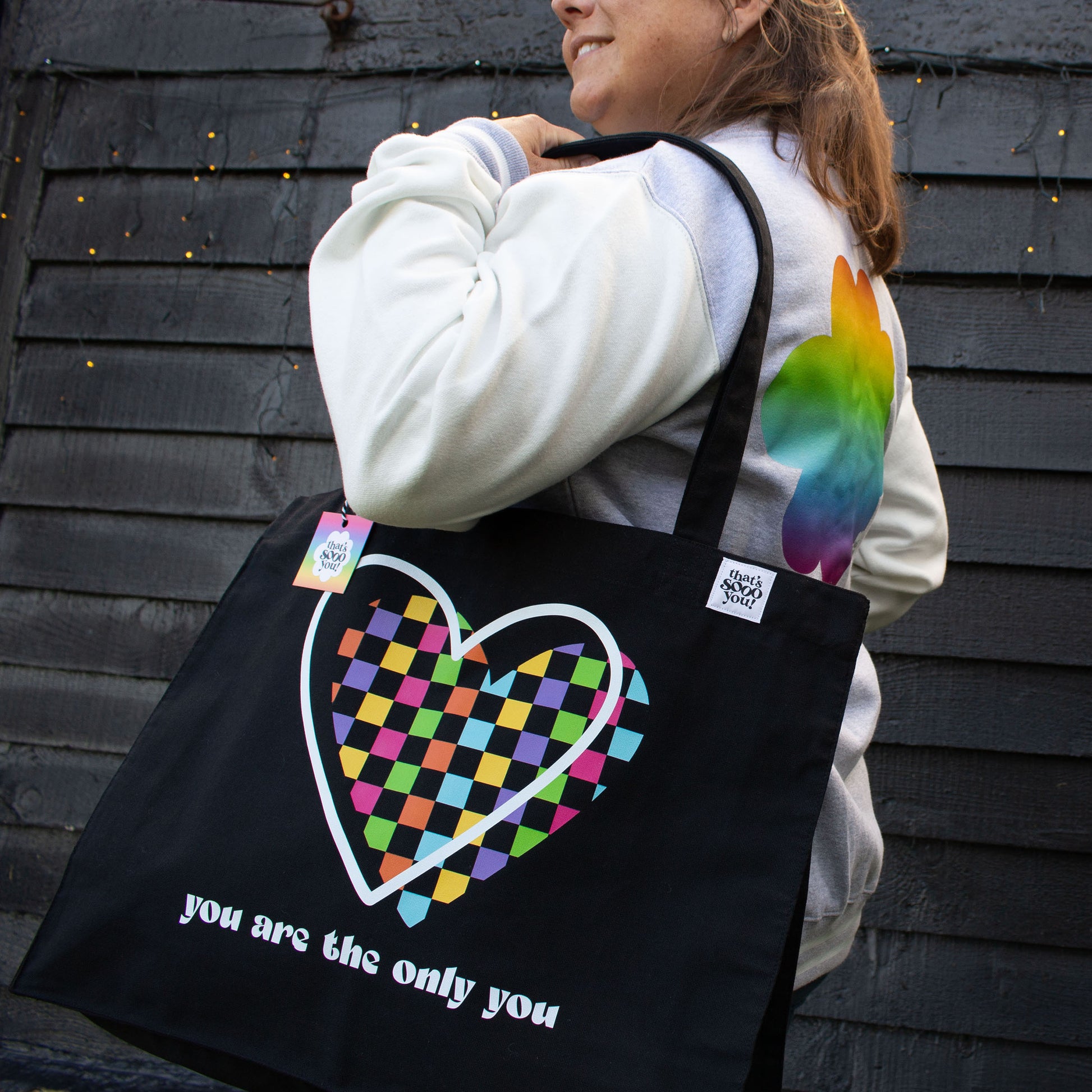 Woman holding black canvas tote bag, That's Sooo You rainbow checkerboard heart You Are The Only You design, she is also wearing a rainbow flower jacket, standing in front of a black wooden wall and facing the camera.