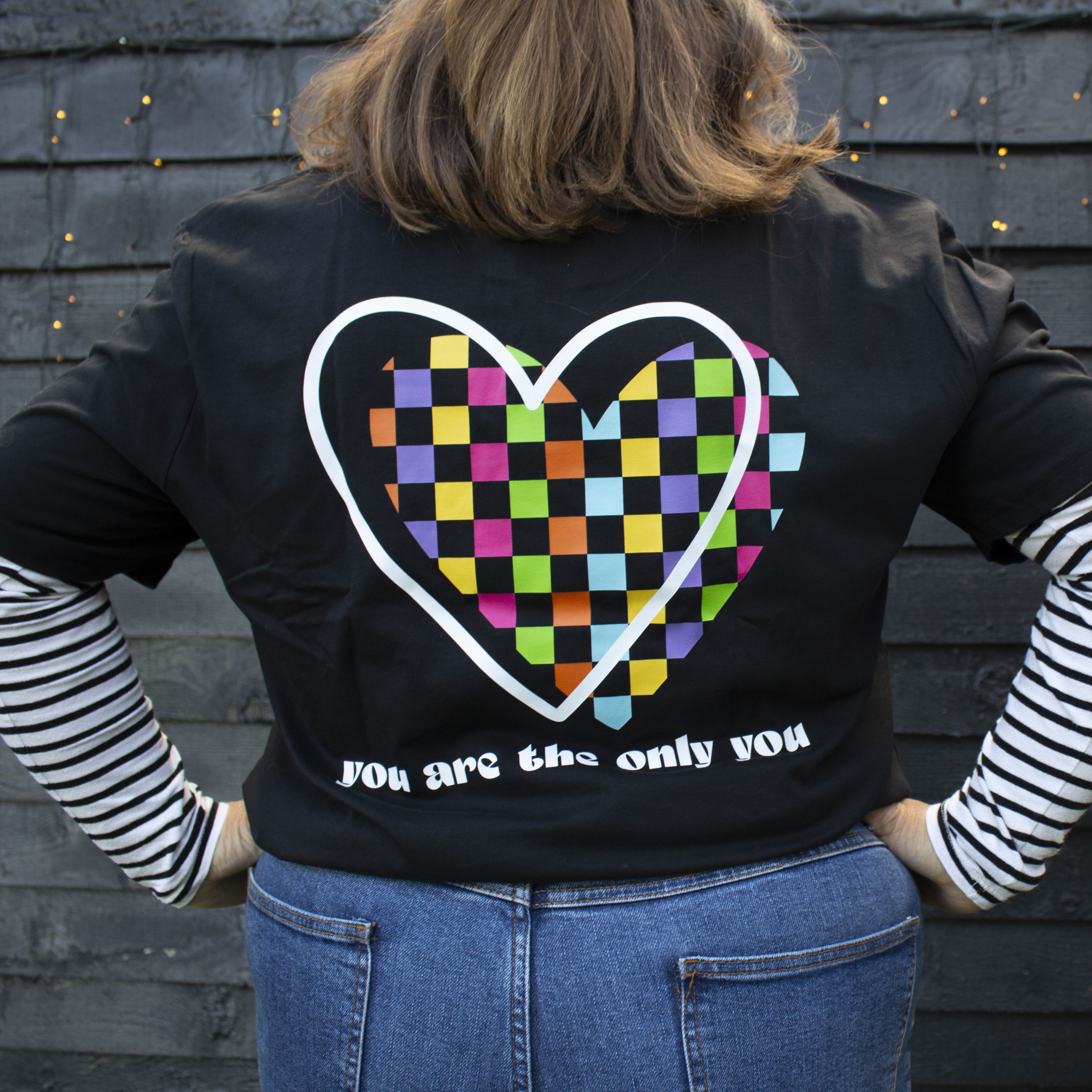 Woman wearing a black That's Sooo You t-shirt with rainbow checkerboard heart on it, it says You Are the Only You, she is facing away from the camera, standing in front of a black wooden wall.