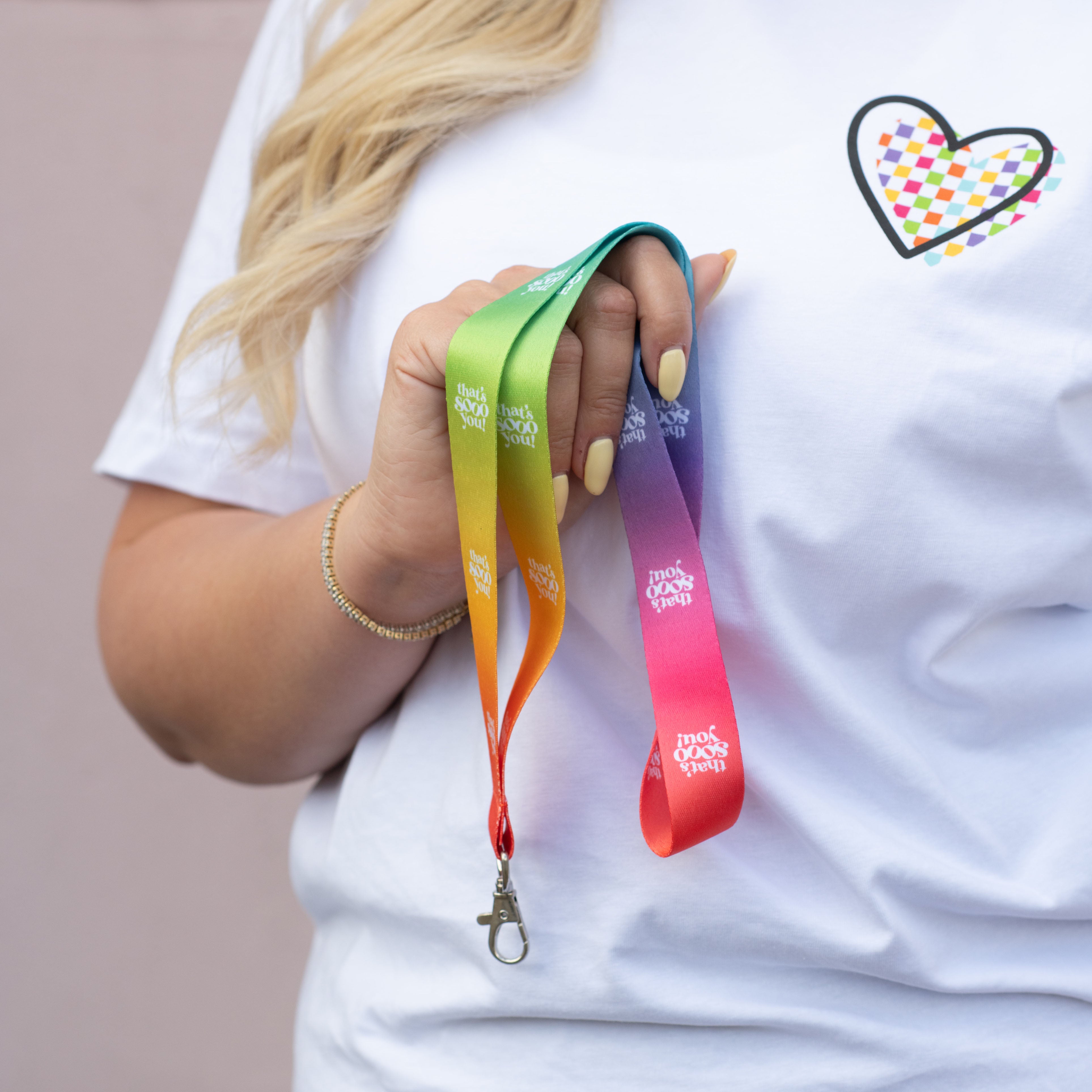 Woman holding TSY rainbow coloured lanyard in her hand, she has pale yellow coloured finger nails, she is wearing a white TSY checkboard heart t-shirt and is standing in front of a pink wall.