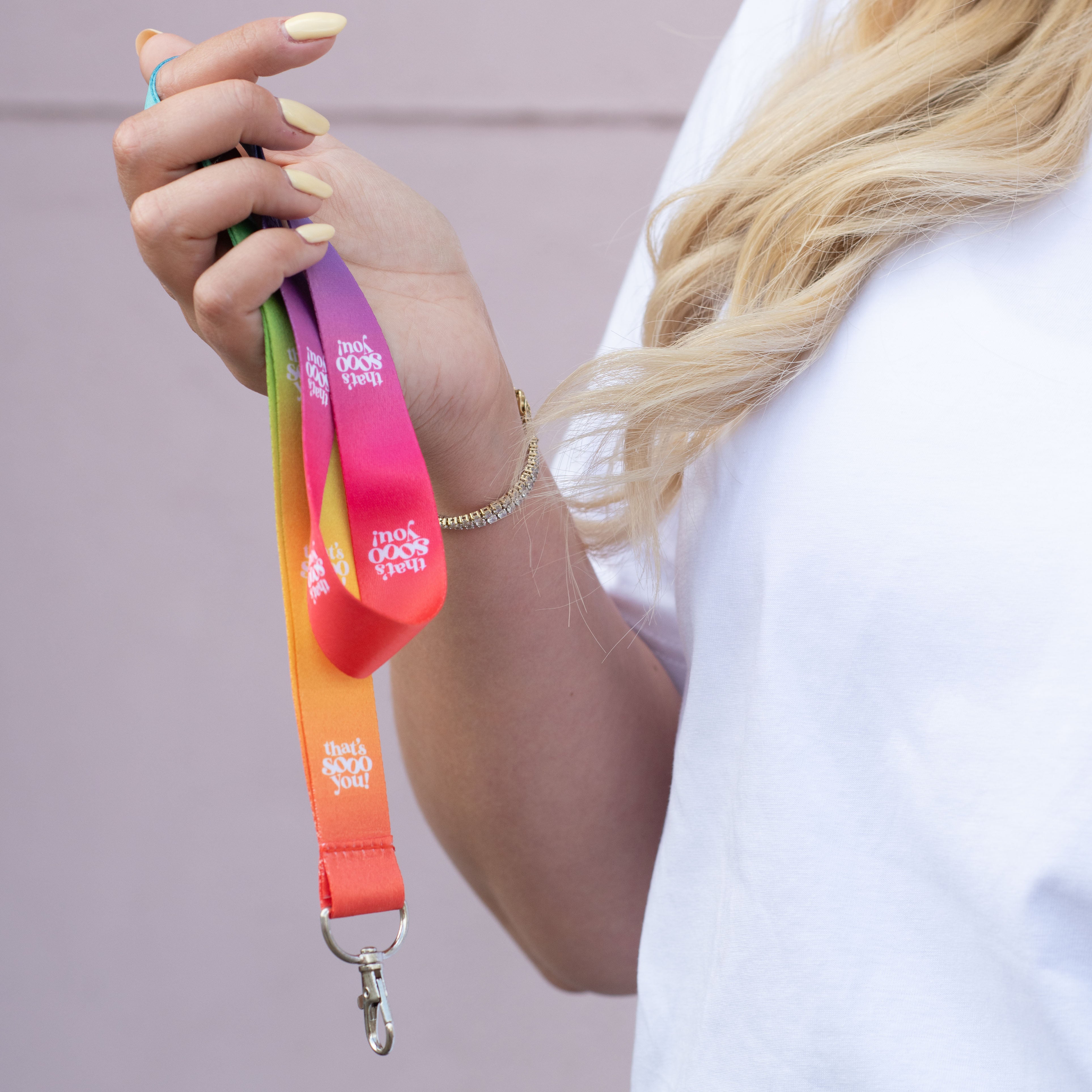 Woman holding TSY rainbow coloured lanyard in her hand, she has pale yellow coloured finger nails, she is wearing a white TSY checkboard heart t-shirt and is standing in front of a pink wall.