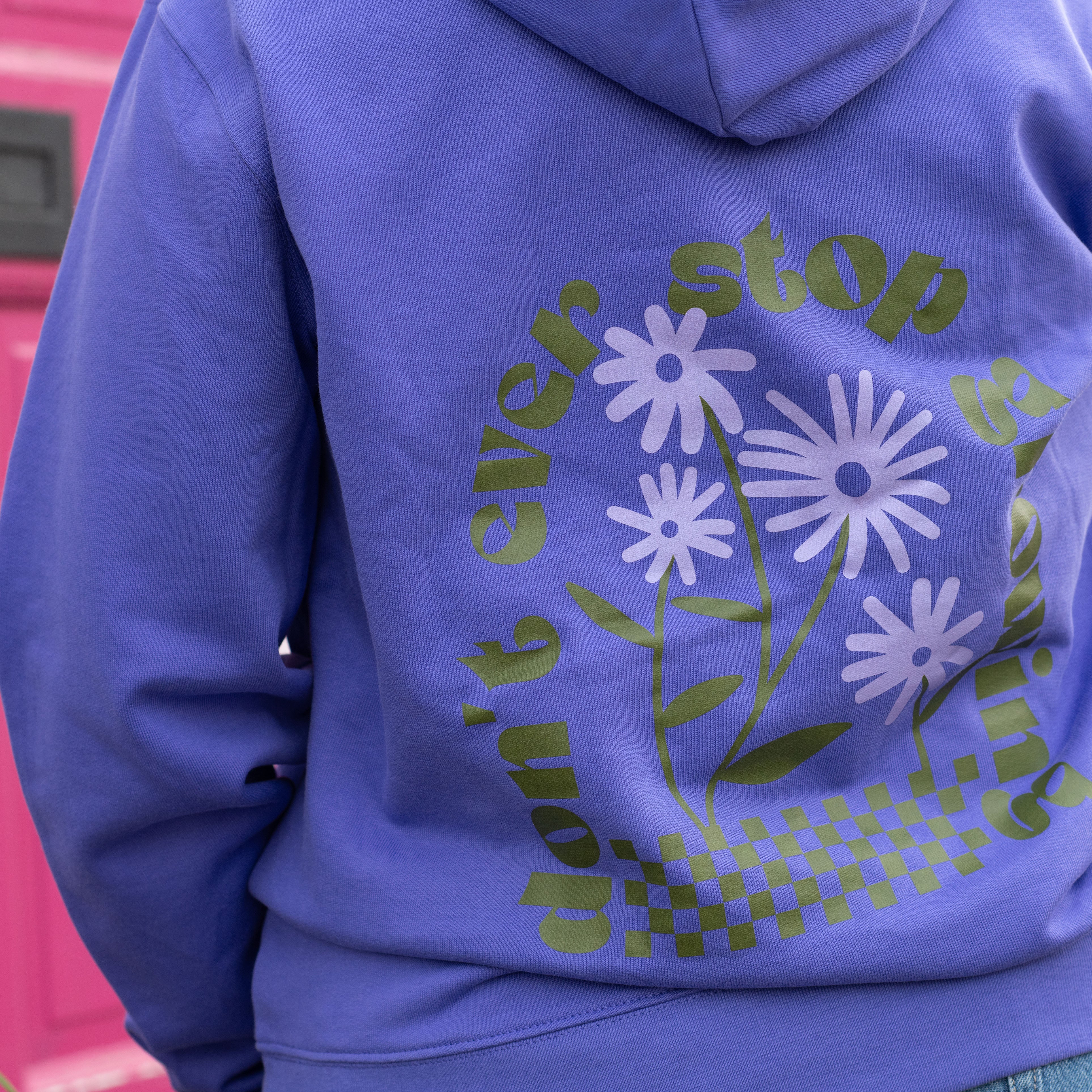 Woman wearing TSY purple hoodie, don't ever stop growing design, with flowers, checkerboard, olive green, lilac print, she is standing in front of a brick wall and she is looking away from the camera.
