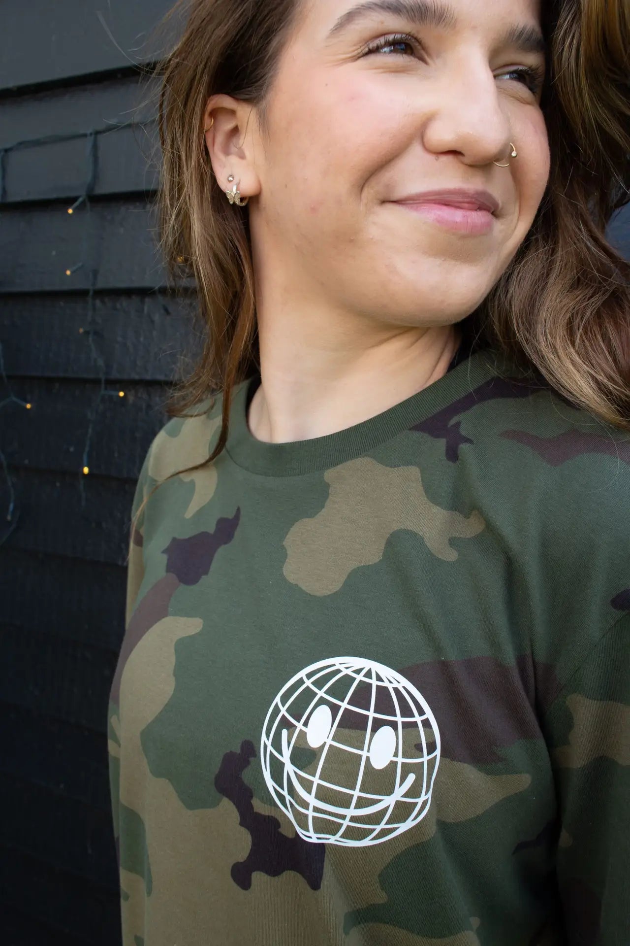 Woman wearing a green camouflage That's Sooo You t-shirt with a white printed, smiley face design, it says Being Weird Feels Right, she is standing in front of a black wooden background with fairy lights, facing the camera.