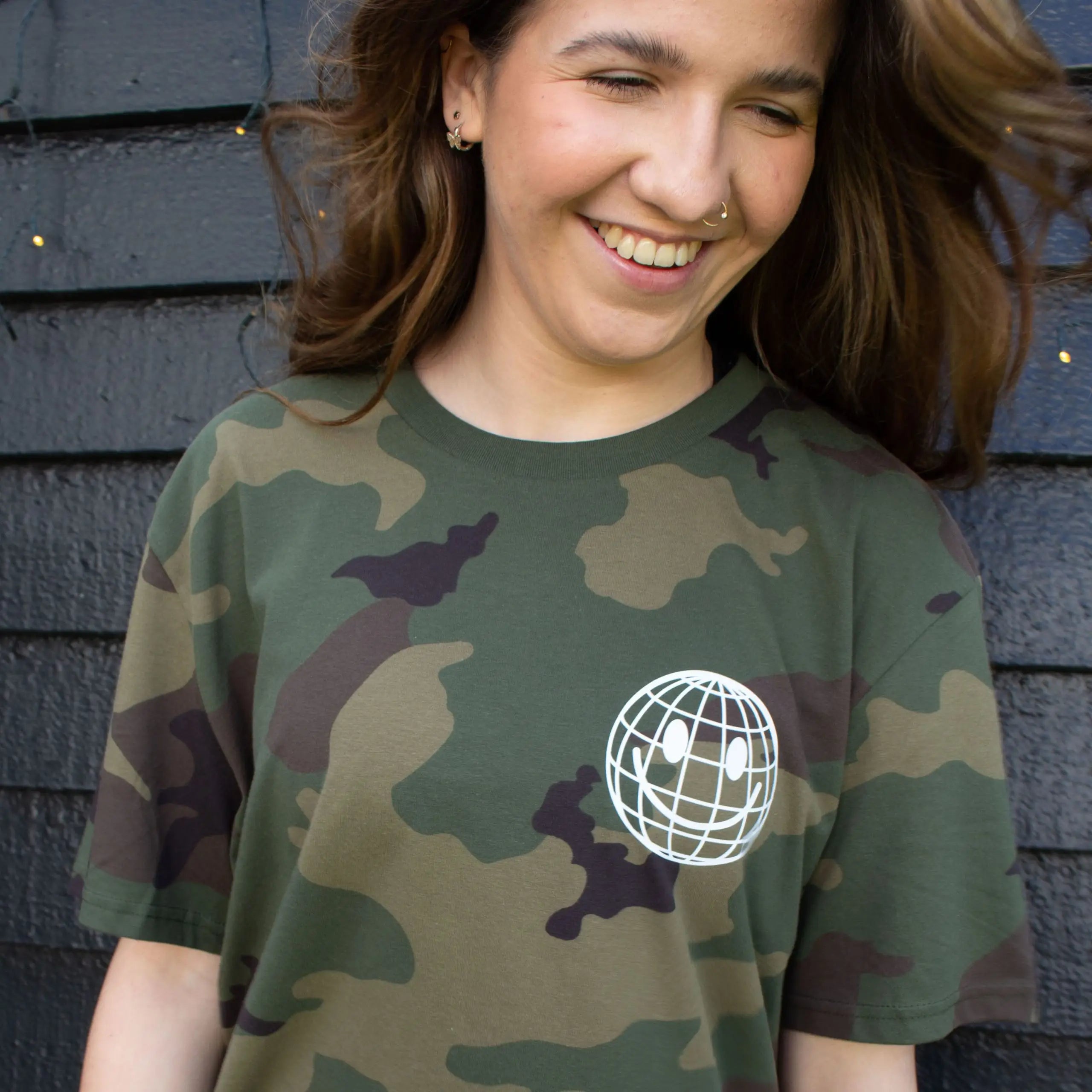 Woman wearing a green camouflage That's Sooo You t-shirt with a white printed, smiley face design, it says Being Weird Feels Right, she is standing in front of a black wooden background with fairy lights, facing the camera and has a big smile.
