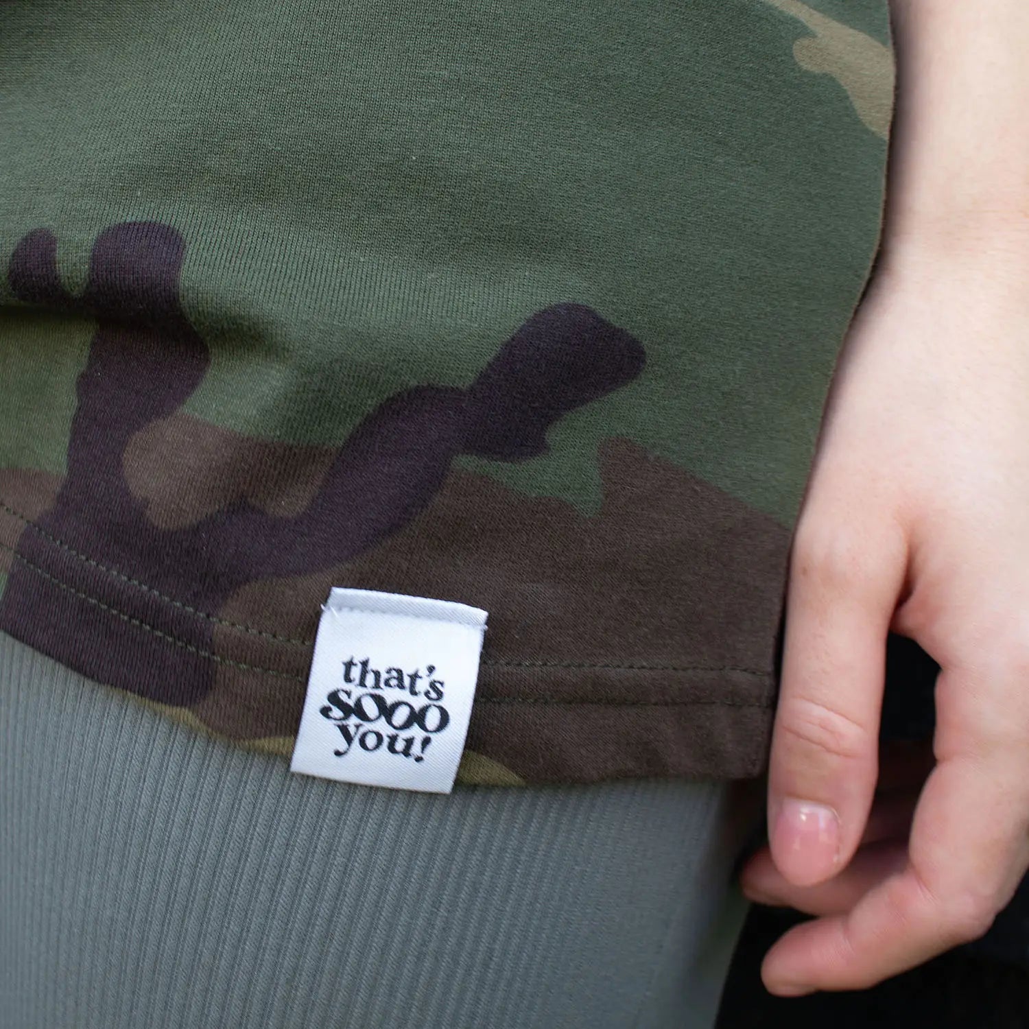 Woman wearing a green camouflage That's Sooo You t-shirt with a white printed, smiley face design, it says Being Weird Feels Right, she is standing in front of a black wooden background with fairy lights, facing the camera, this is a close up of the hand-sewn TSY label.