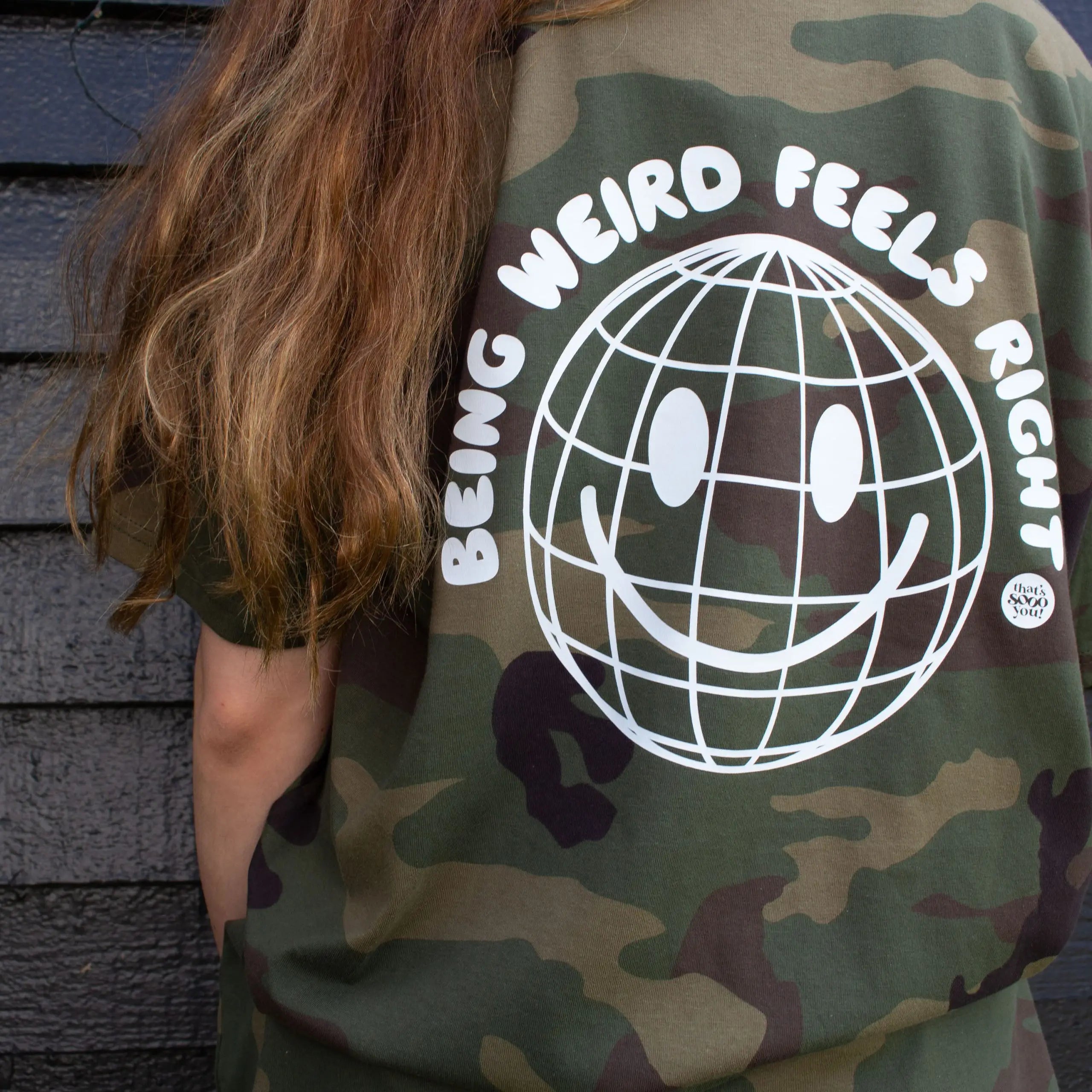 Woman wearing a green camouflage That's Sooo You t-shirt with a white printed, smiley face design, it says Being Weird Feels Right, she is standing in front of a black wooden background with fairy lights, facing away from the camera.