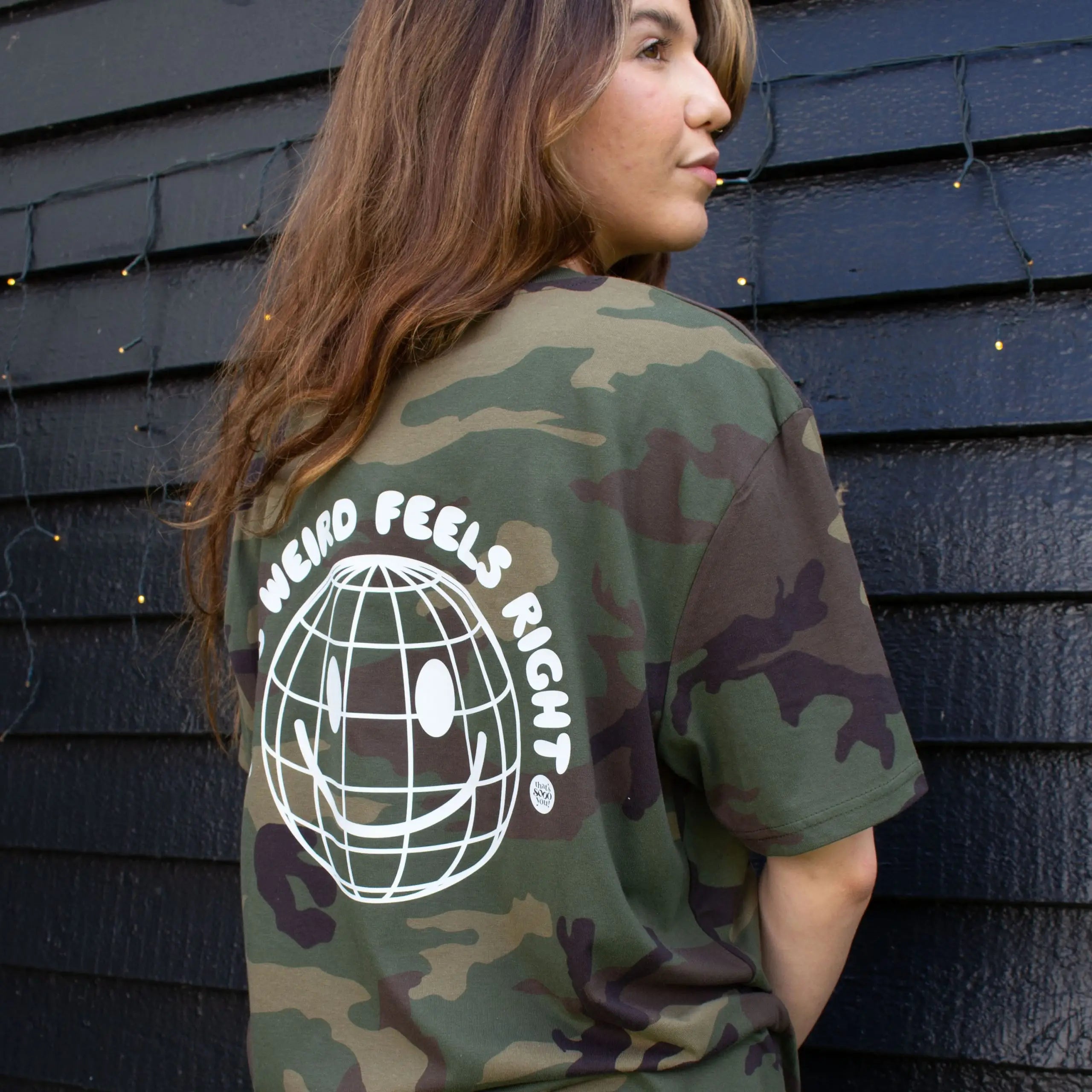 Woman wearing a green camouflage That's Sooo You t-shirt with a white printed, smiley face design, it says Being Weird Feels Right, she is standing in front of a black wooden background with fairy lights, facing away from the camera.