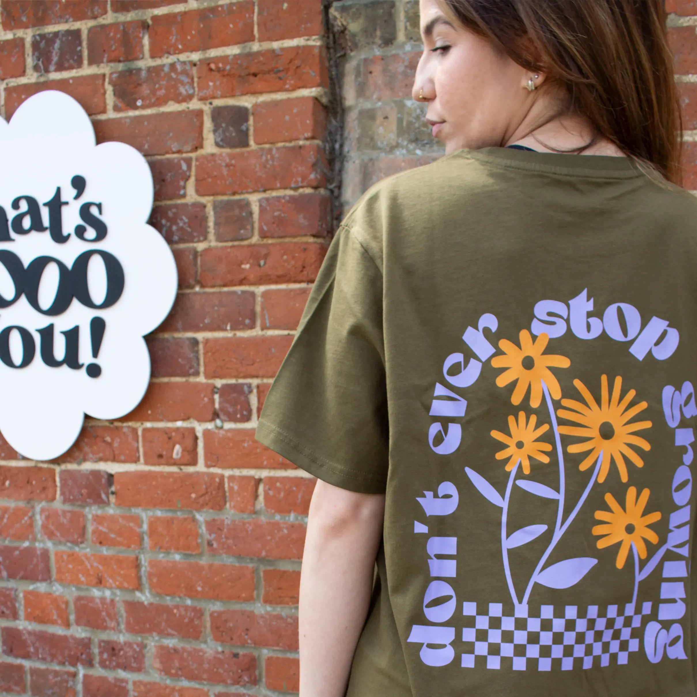 Woman wearing olive green That's Sooo You t-shirt with orange and lilac printed, Don't Ever Stop Growing, she is standing in front of a brick background, with a TSY sign, facing away from the camera.