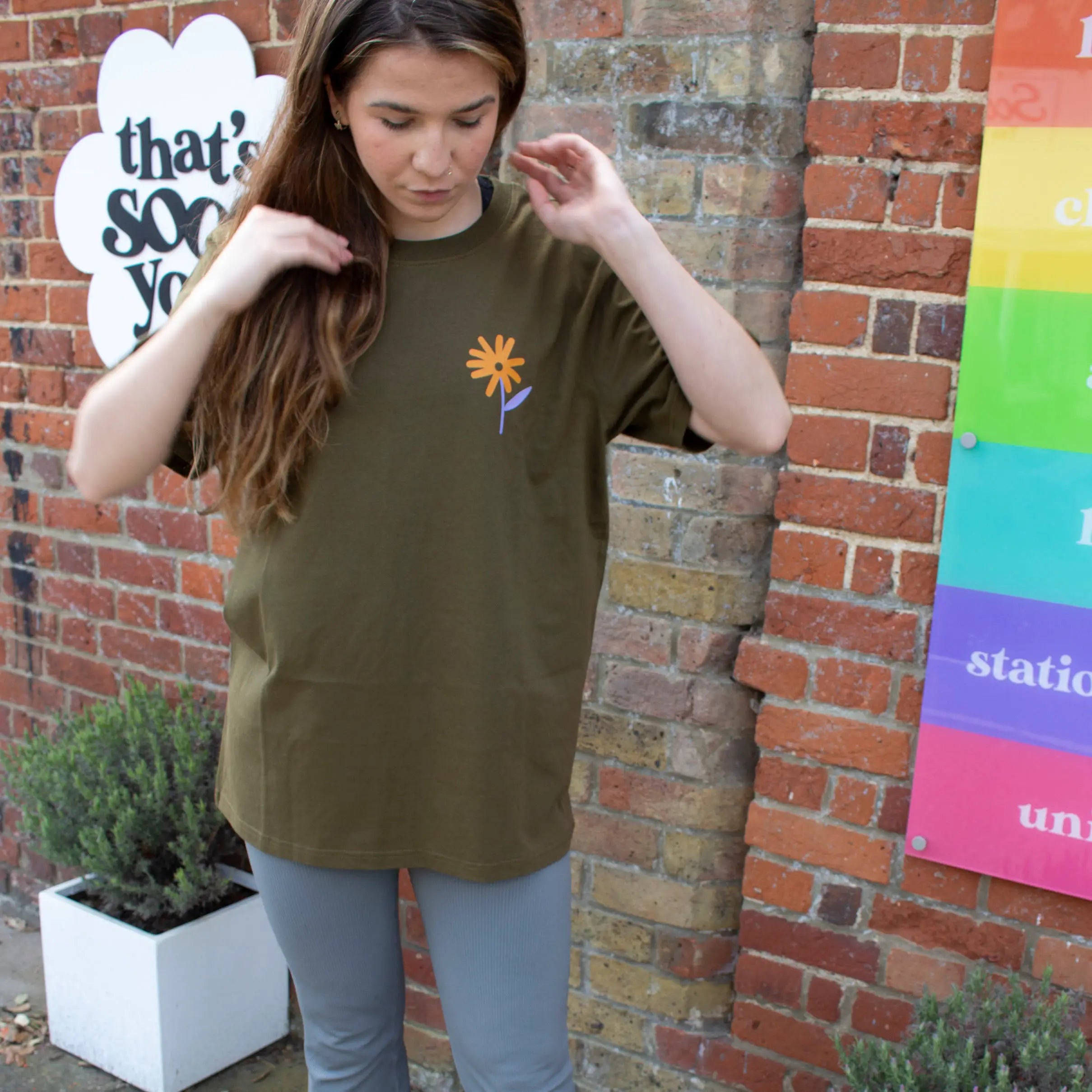 Woman wearing olive green That's Sooo You t-shirt with orange and lilac printed, Don't Ever Stop Growing, she is standing in front of a brick background, with a TSY sign, facing the camera.