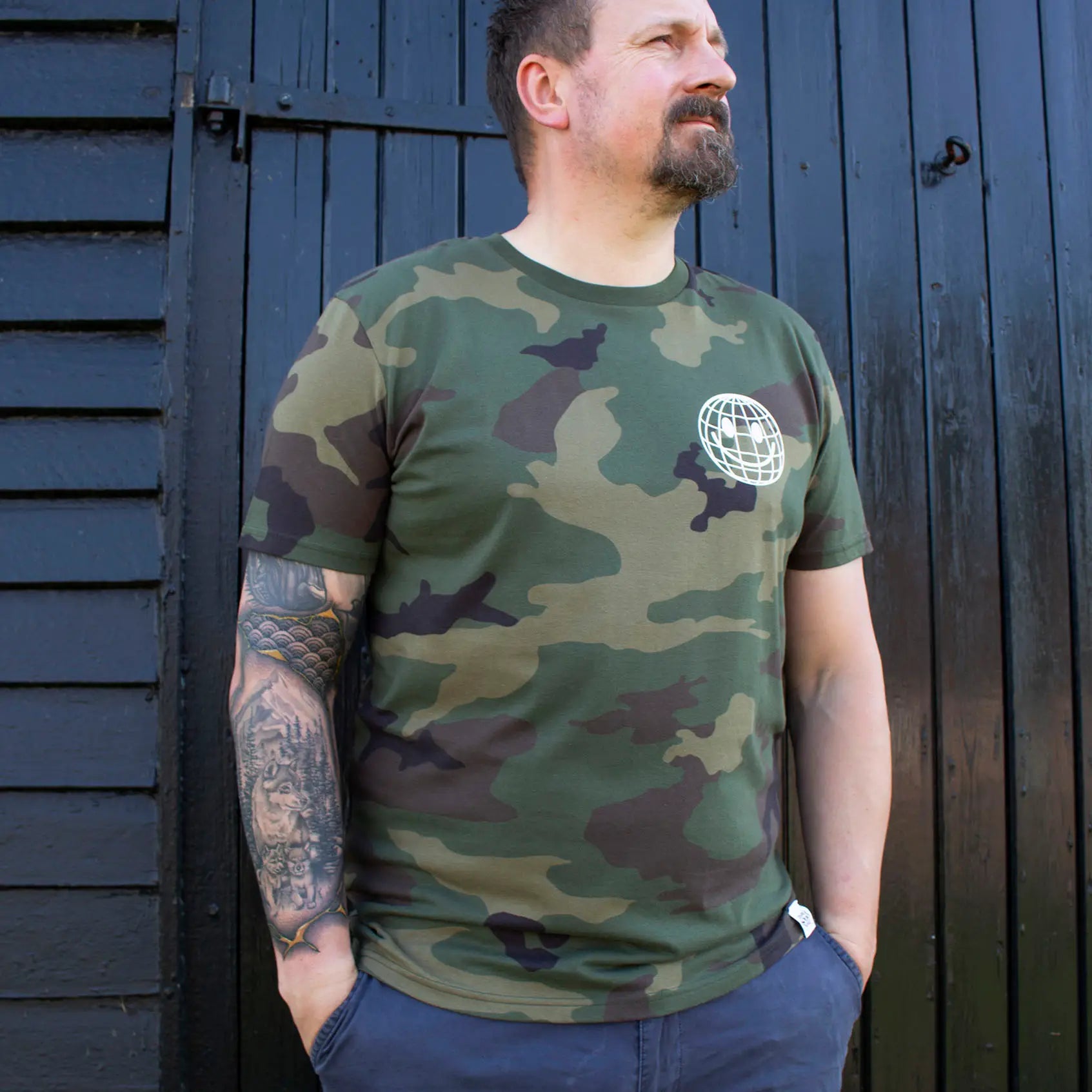 Man wearing a green camouflage That's Sooo You t-shirt with a white printed, smiley face design, it says Being Weird Feels Right, he is standing in front of a black wooden background, facing the camera.