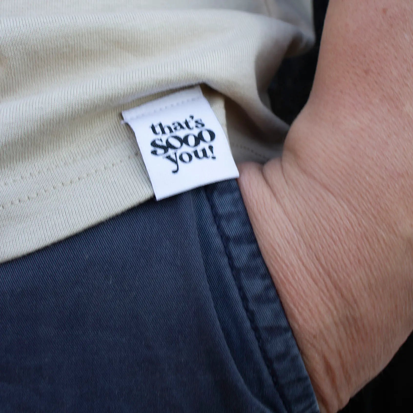 Man wearing beige That's Sooo You t-shirt with ollive printed, smiley face design, it says Being Weird Feels Right, he is standing in front of a black wooden background with fairy lights, facing the camera, this is a close up of his hand in his pocket with a hand-sewn TSY label.