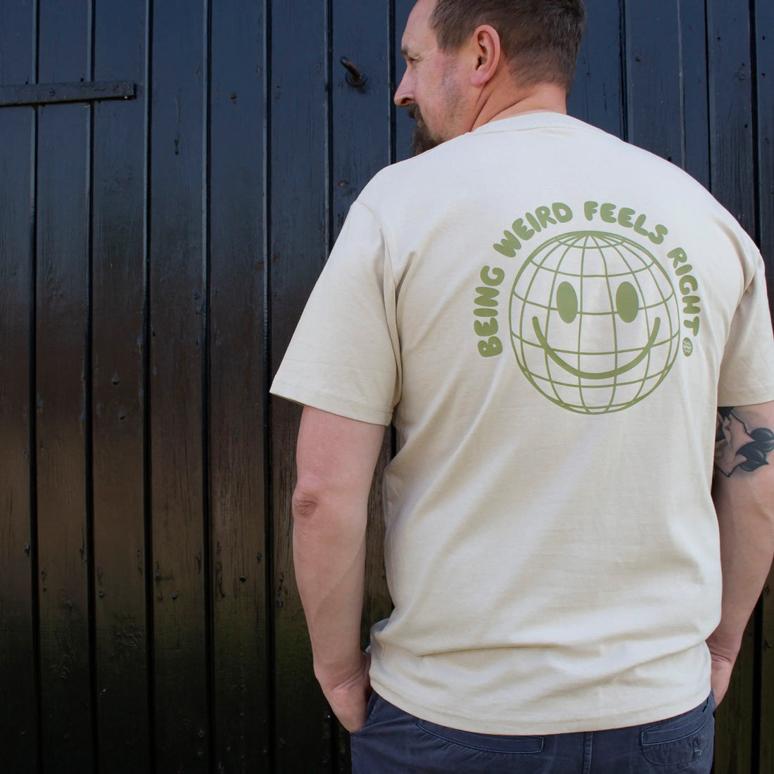 Man wearing beige That's Sooo You t-shirt with ollive printed, smiley face design, it says Being Weird Feels Right, he is standing in front of a black wooden background, facing away from the camera.