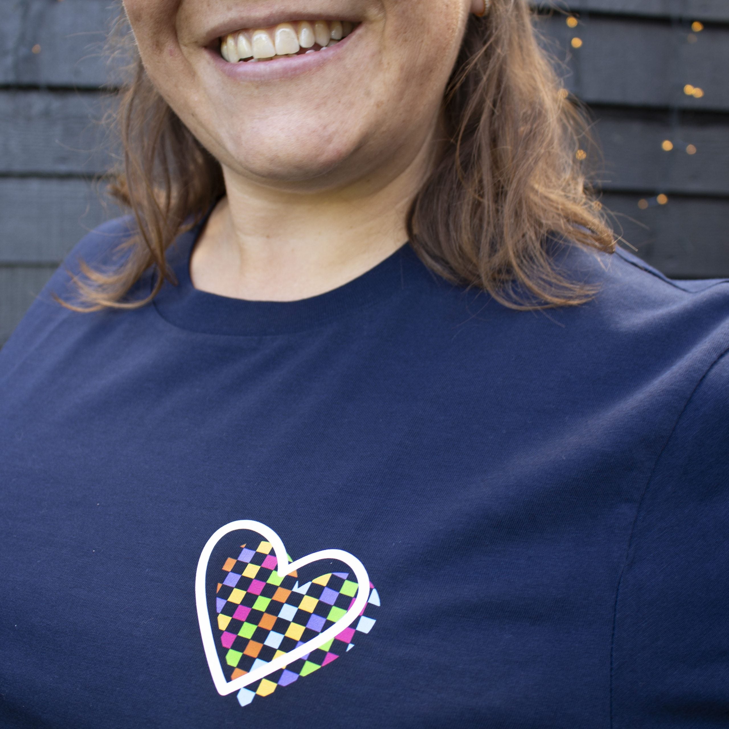 Woman wearing navy That's Sooo You t-shirt with rainbow checkerboard heart on it, it says You Are the Only You, she is smiling at the camera, standing in front of a black wooden wall.