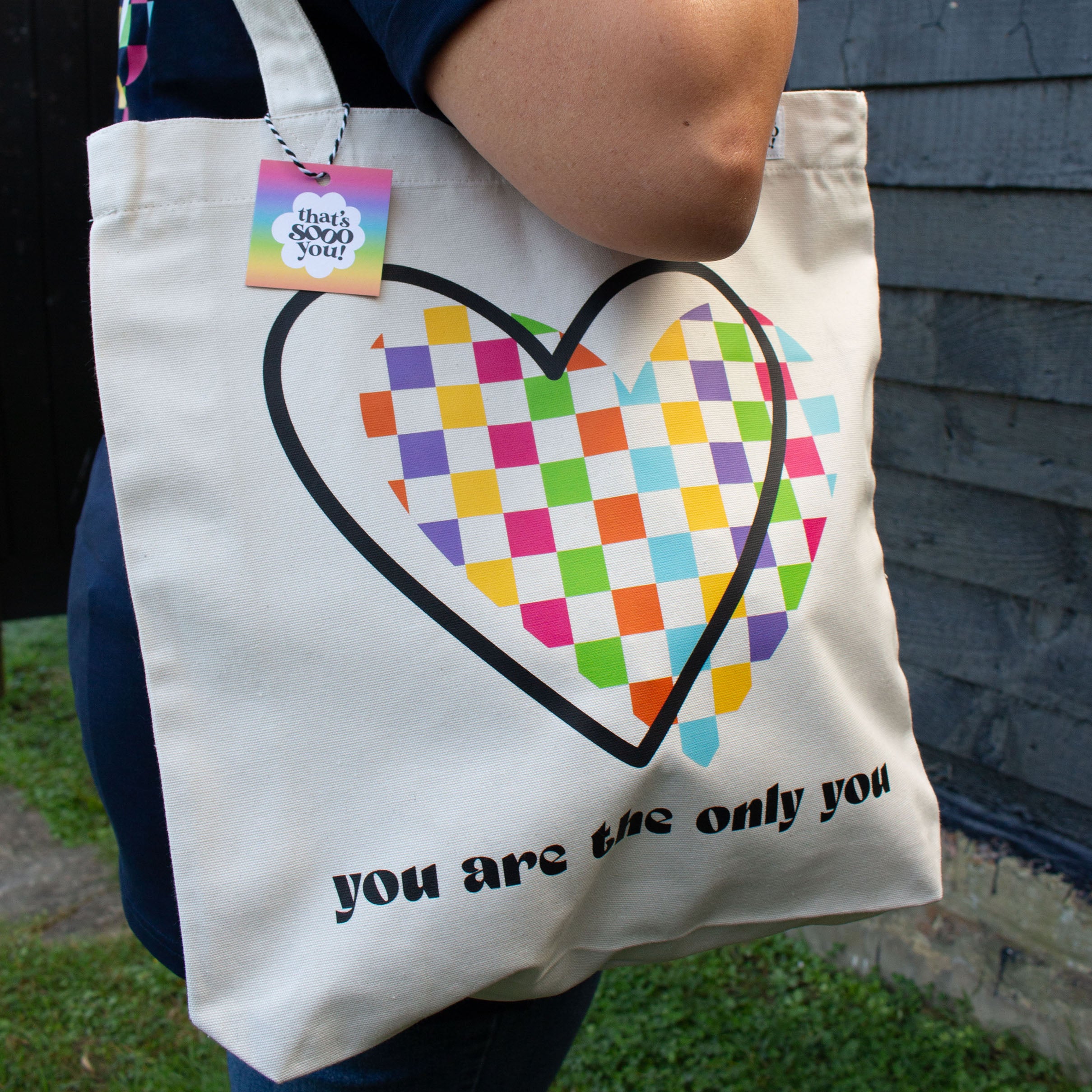 Woman holding cream natural canvas tote bag, That's Sooo You rainbow checkerboard heart You Are The Only You design, she is standing in front of a black wooden wall and facing the camera.