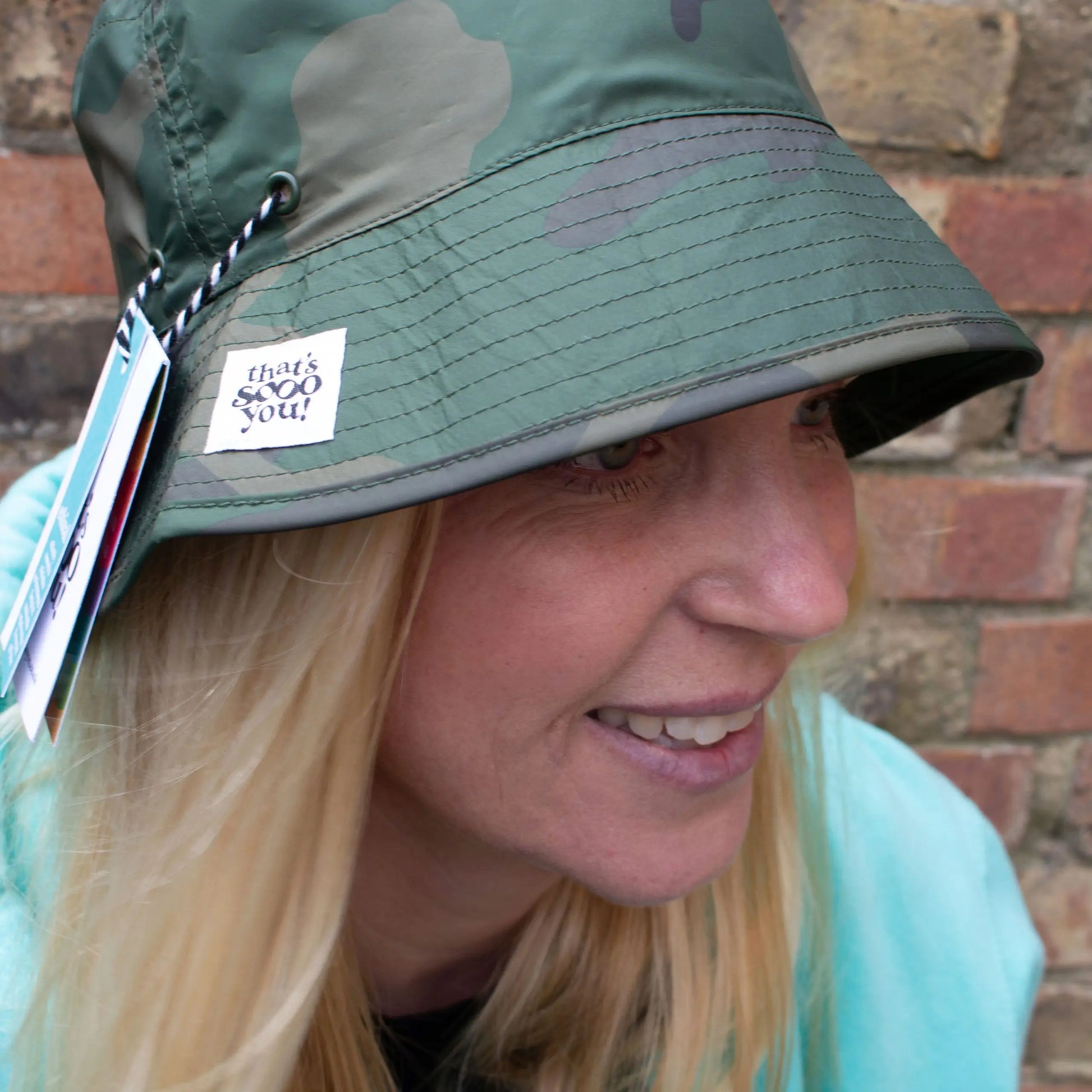 Woman wearing green camouflage bucket hat with That's Sooo You hand-sewn label on it, she is standing in front of a brick wall and facing the camera.