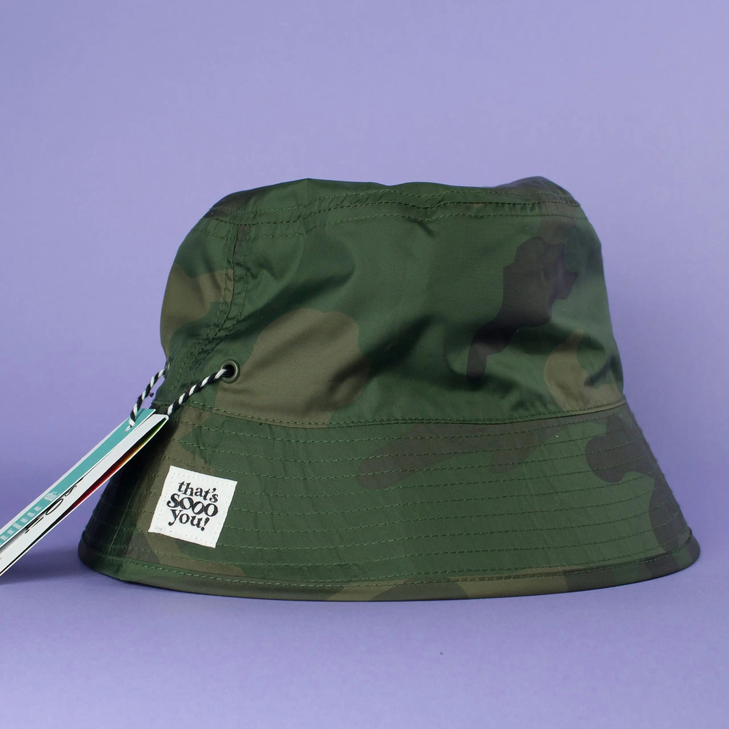 Green camouflage bucket hat with That's Sooo You hand-sewn label on it, photographed on a lilac purple background.