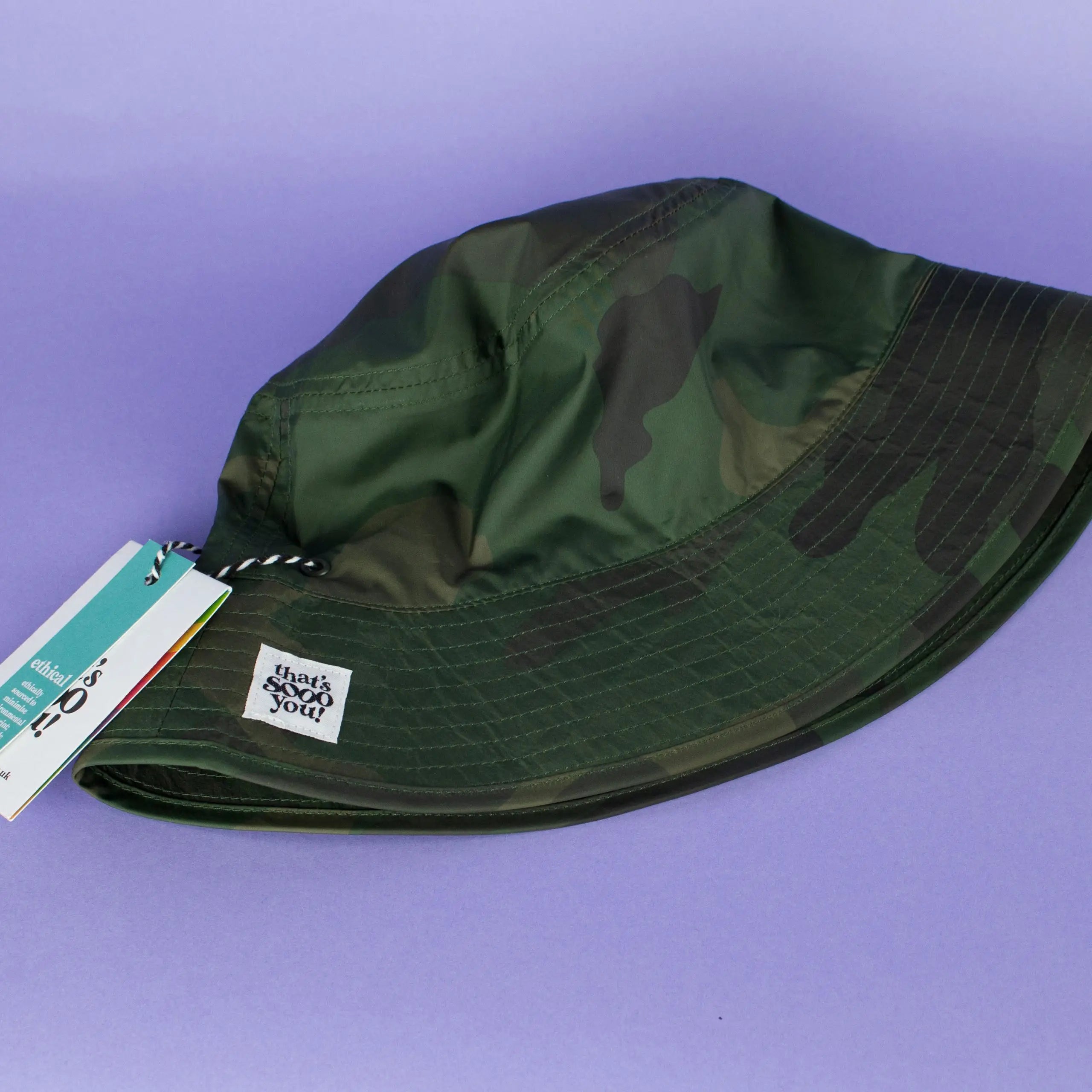 Green camouflage bucket hat with That's Sooo You hand-sewn label on it, photographed on a lilac purple background.