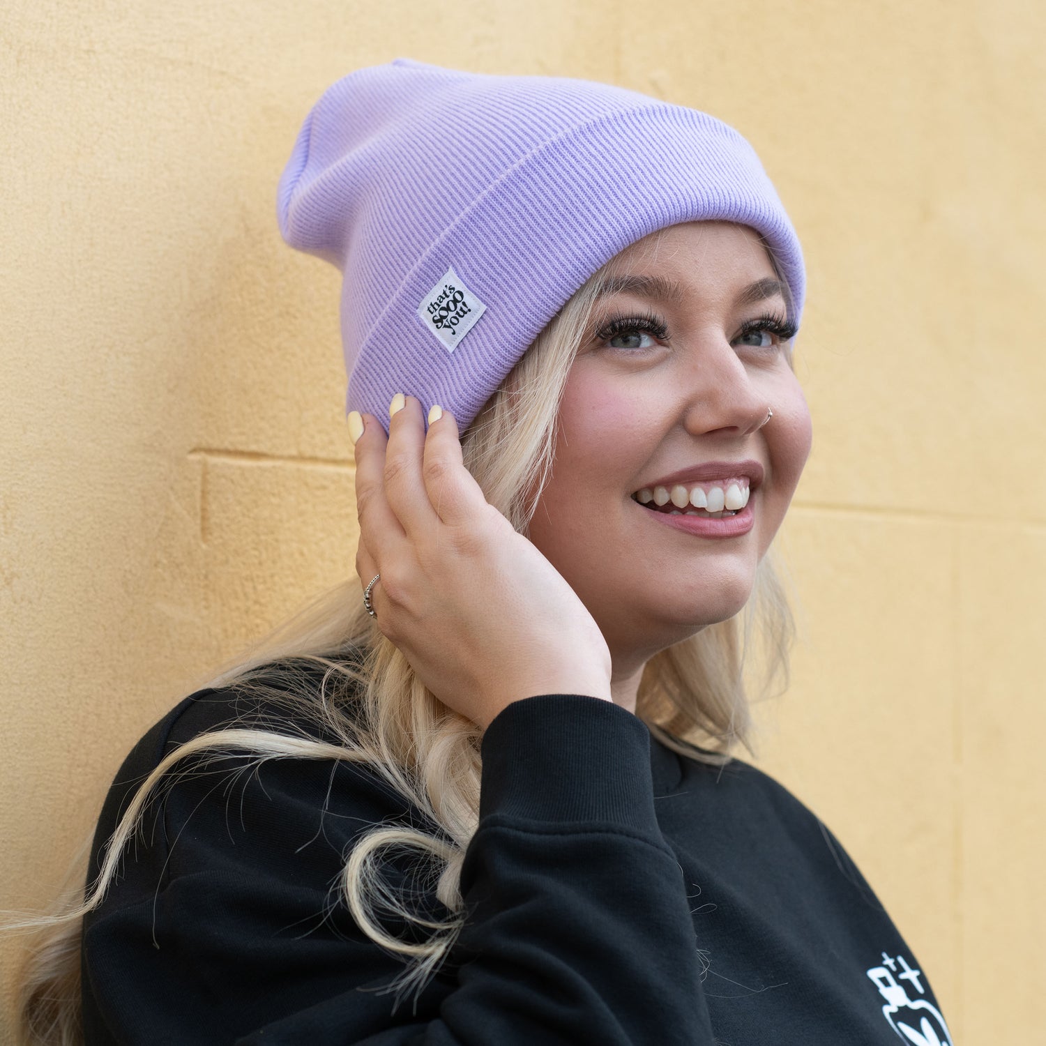 Woman is standing in front of a pale yellow coloured wall, she is wearing a black TSY sweatshirt dress, she is also wearing a lilac TSY beanie hat, she is smiling and touching her hat with her hand, she is looking away from the camera.