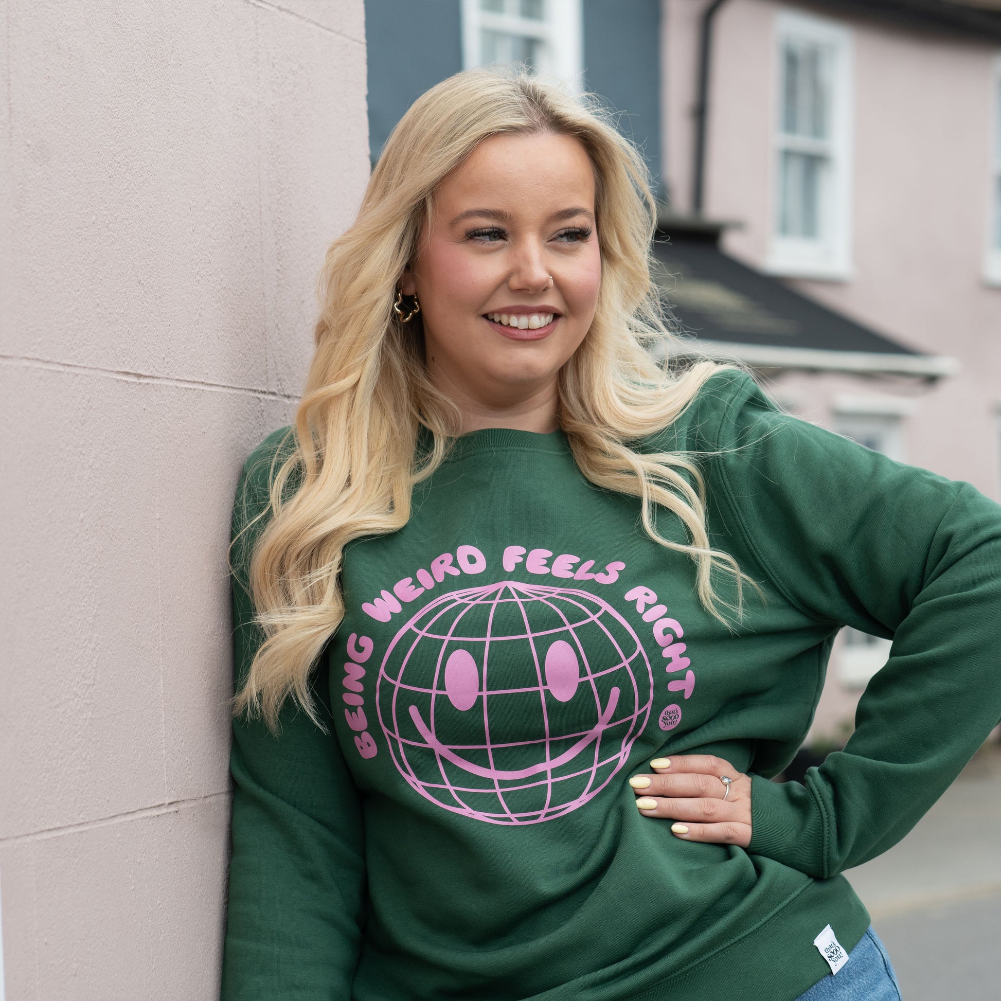 Model wearing TSY Keep Being Weird green sweatshirt with pink printing, standing in front of a pink wall and white window, she is smiling and looking away from the camera and has her hand on her hip.