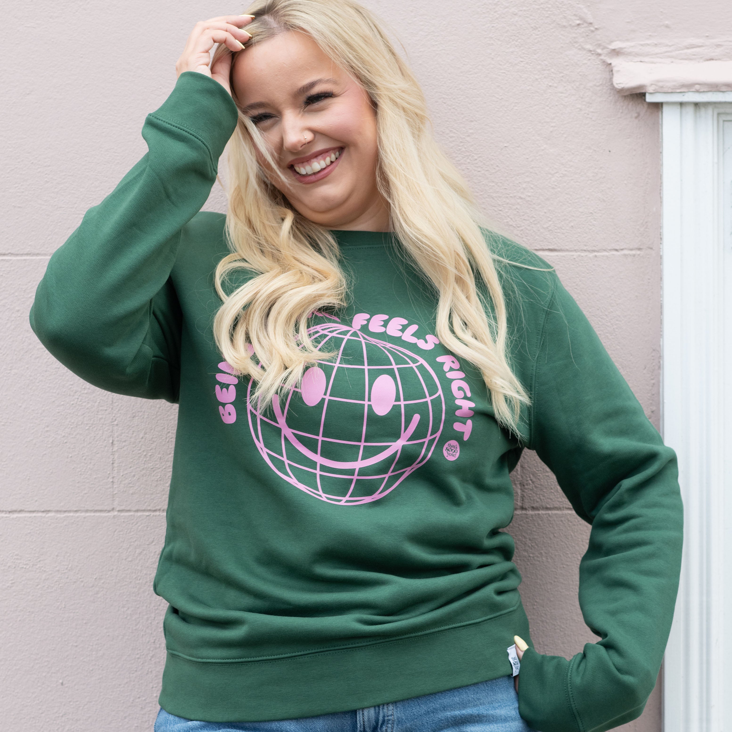 Model wearing TSY Keep Being Weird green sweatshirt with pink printing, standing in front of a pink wall and white window, she is smiling and looking away from the camera with a hand in her hair.