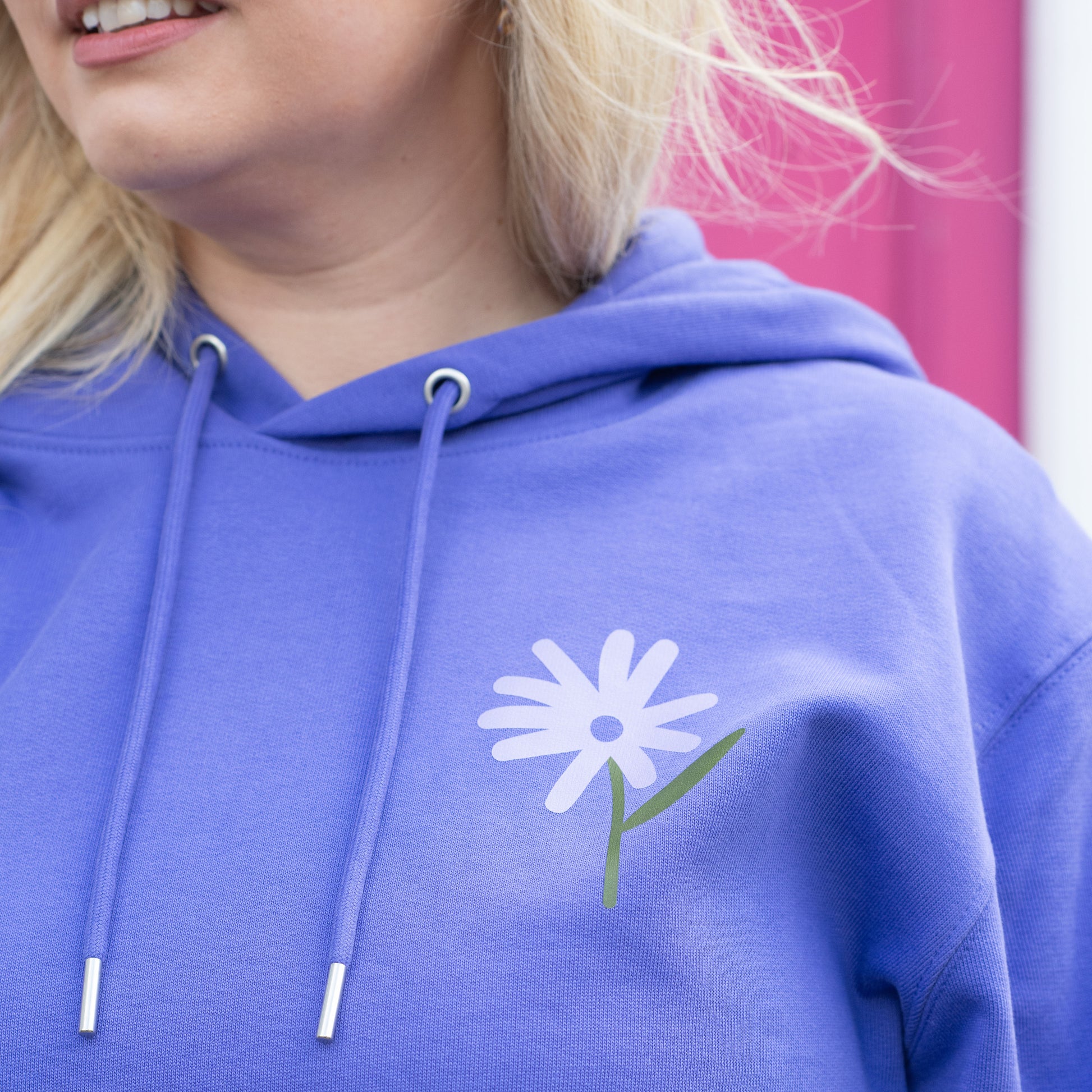 Woman wearing TSY purple hoodie, don't ever stop growing design, with flowers, checkerboard, olive green, lilac print, she is standing in front of a brick wall and she is smiling looking away from the camera.